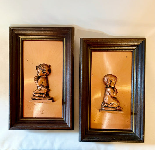 Two framed copper sculptures of children on a white wall.